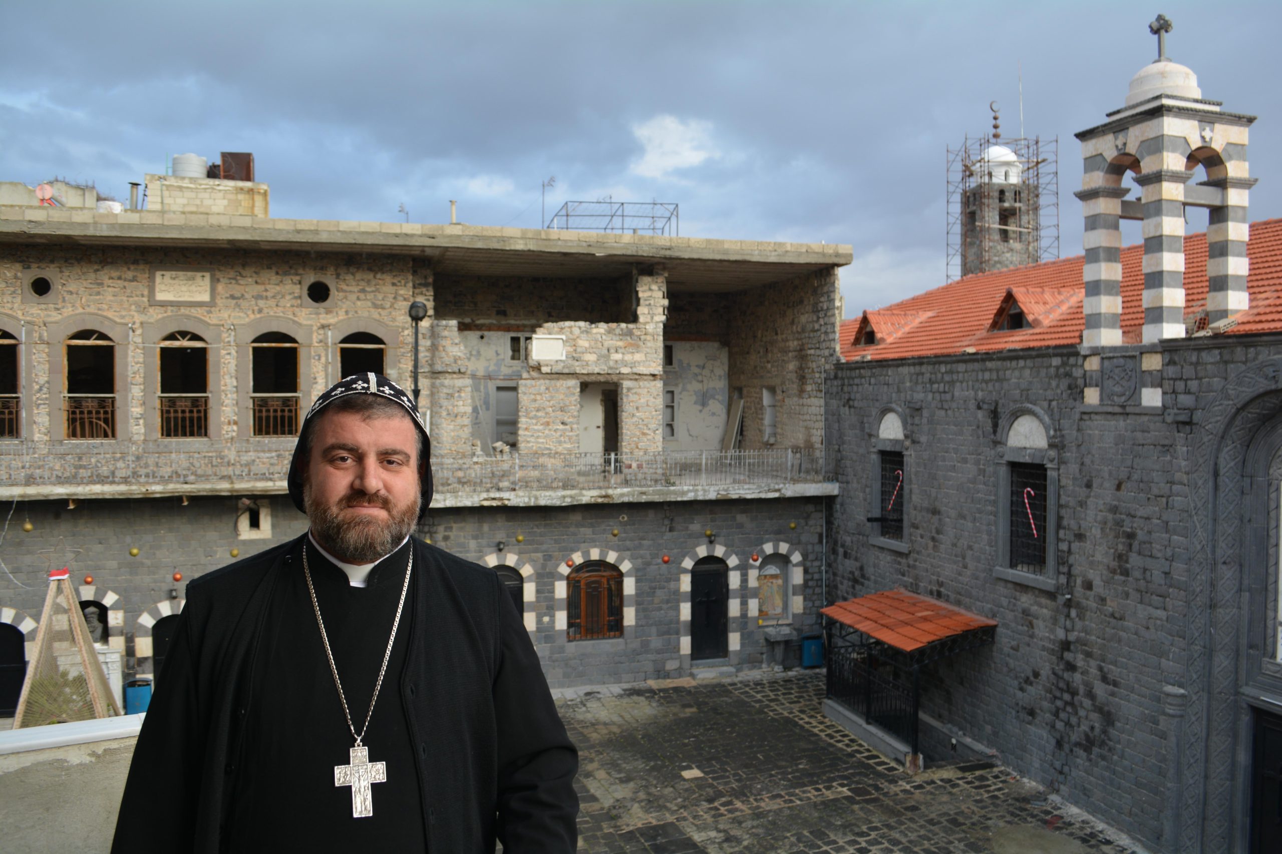 Syriac Orthodox Archbishop Selwanos Petros Al-Nemeh of Homs and Hama, with Saint Mary Church of the Holy Belt in the background (© Aid to the Church in Need)