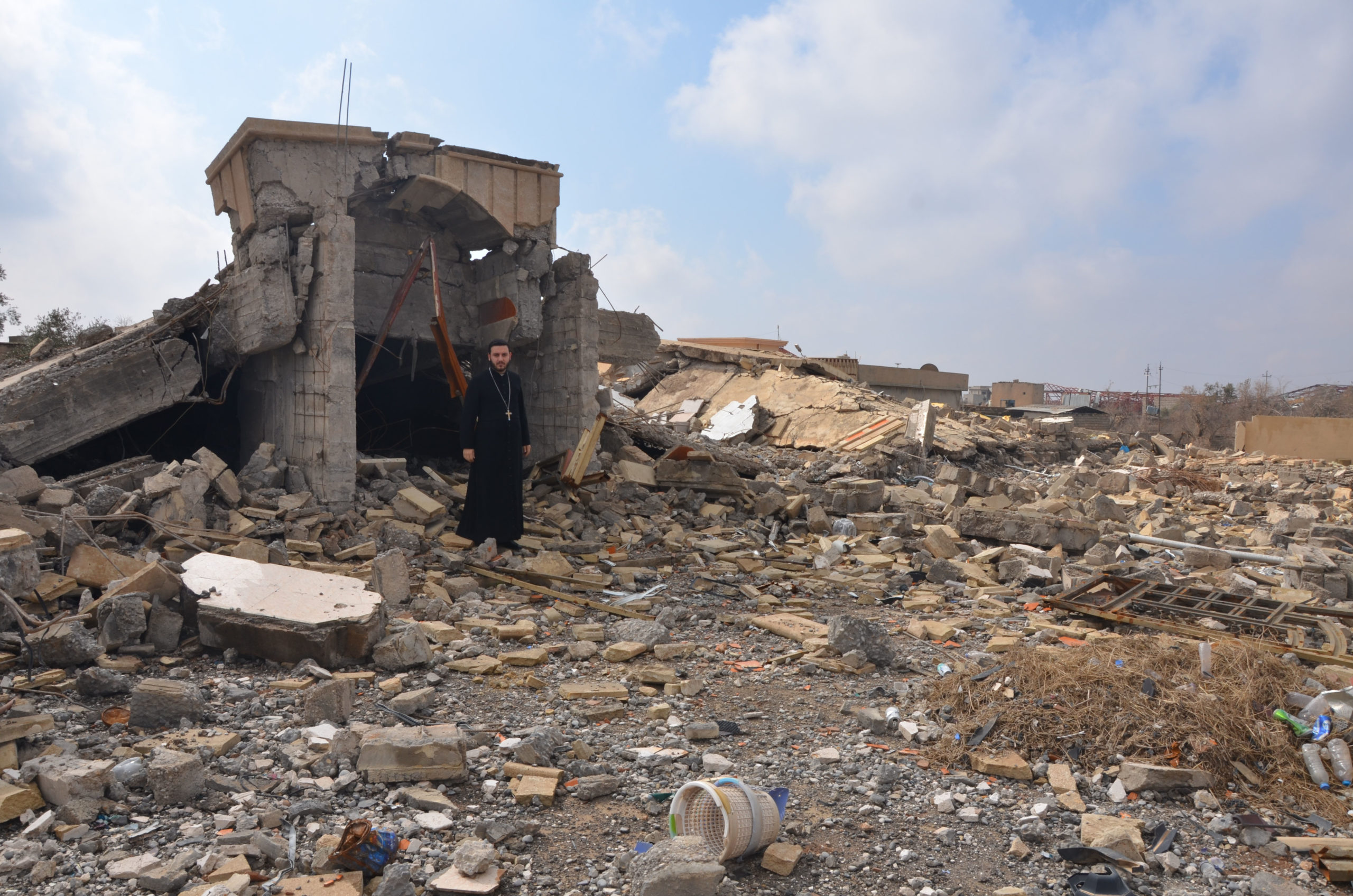 With image of a priest standing amid the ruins of a building in Bashiqa destroyed during Daesh (ISIS)’s occupation of the town (Image © Aid to the Church in Need)